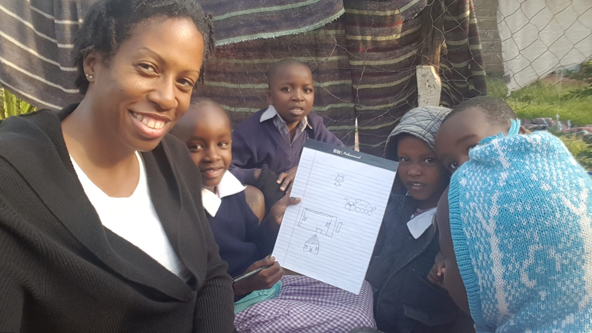 Kennesha seated outdoors with young students in Kenya as they gather around a notebook showing a child’s drawings during the 2018 mission.