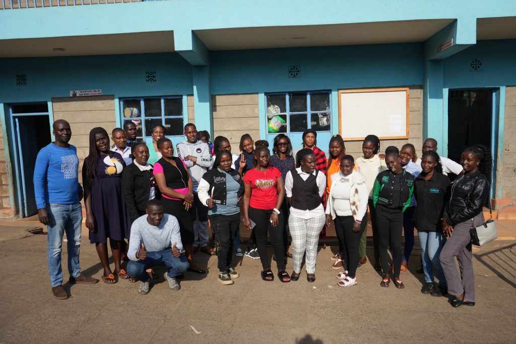 A large group of adults pose together outside a blue building, with several people smiling and giving thumbs-up gestures.
