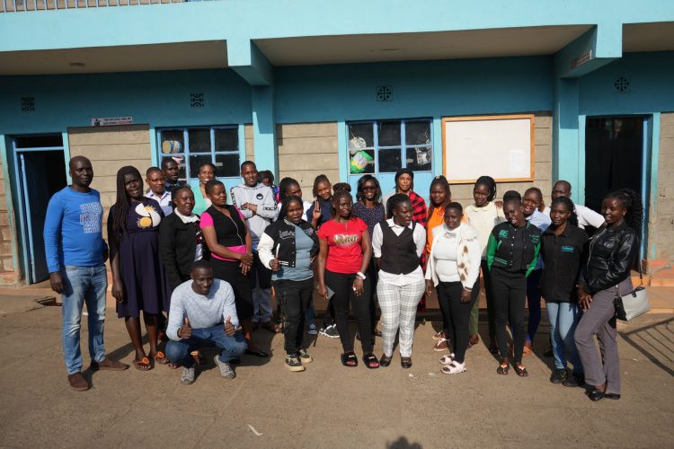 A large group of adults pose together outside a blue building, with several people smiling and giving thumbs-up gestures.
