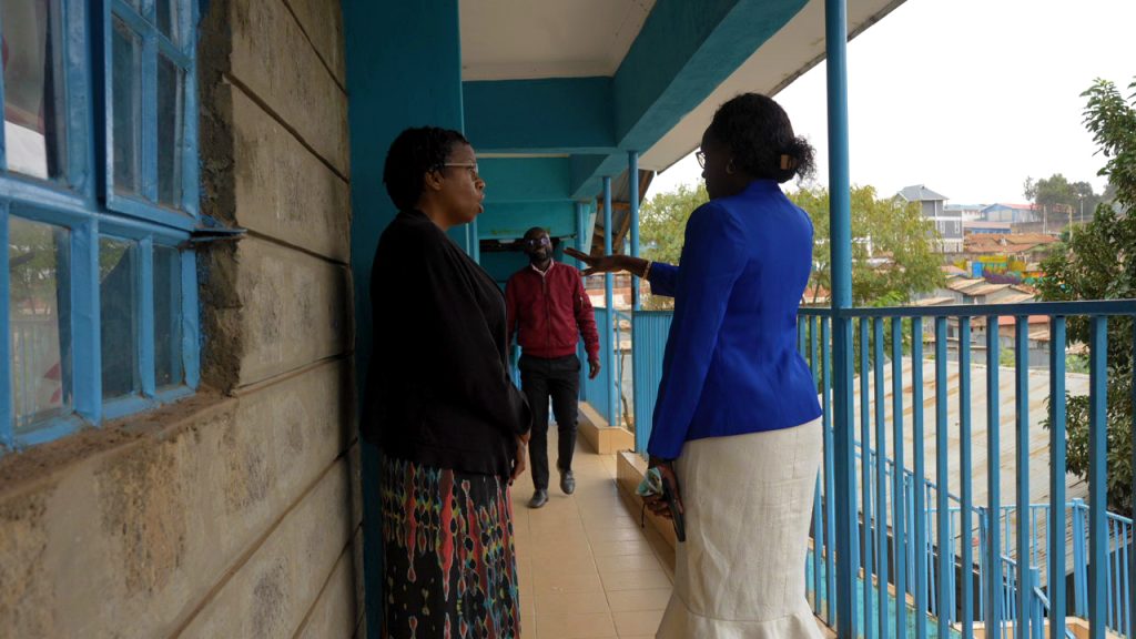 Two women converse on a blue-railed outdoor school corridor while a man walks toward them in the background.