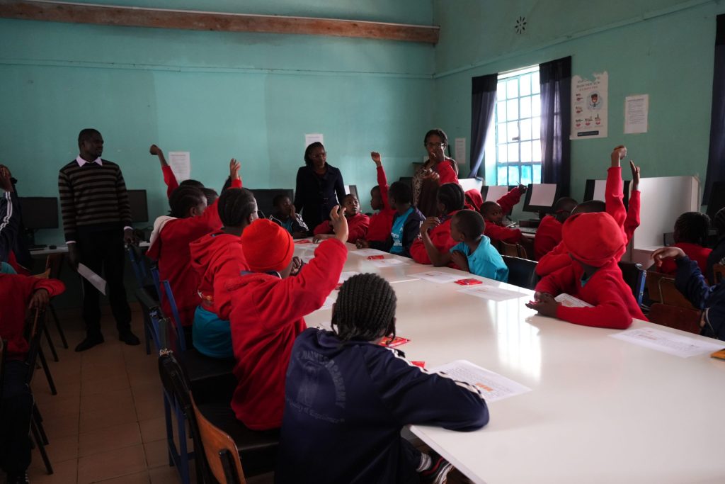 Many schoolchildren seated around a large white table raise their hands while instructors stand at the front of the computer lab.