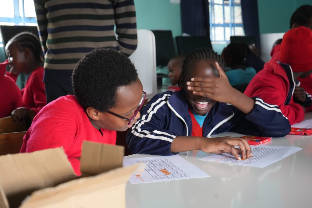 An adult assists a laughing schoolgirl who covers her eyes while working with a printed keyboard exercise.