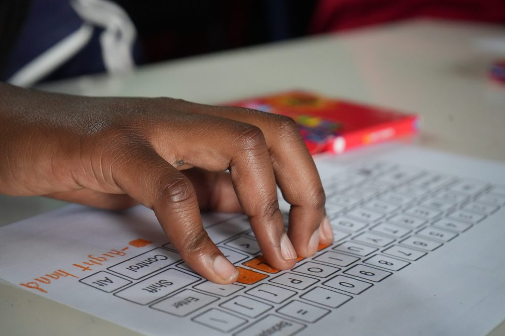Close-up of a hand pressing orange-highlighted keys on a printed computer keyboard worksheet.