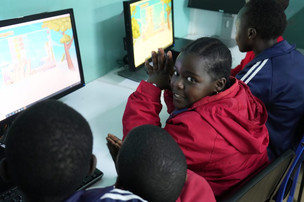 A schoolgirl in a red jacket turns toward the camera while seated with classmates at desktop computers.