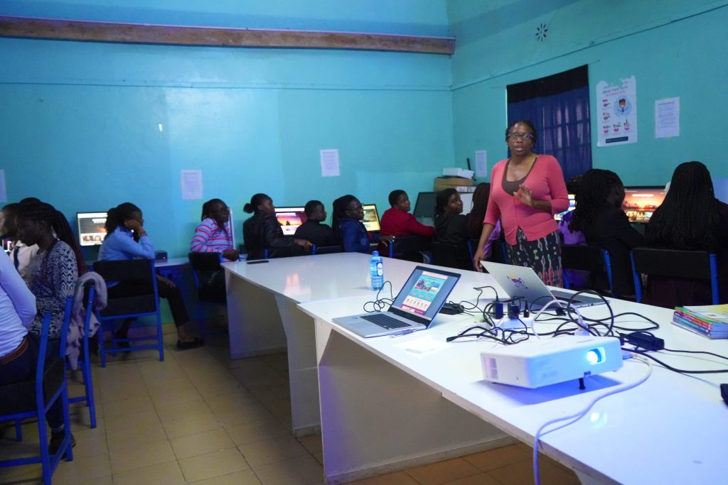 A woman stands addressing adult learners in a computer lab with laptops, a projector, and desktop workstations.