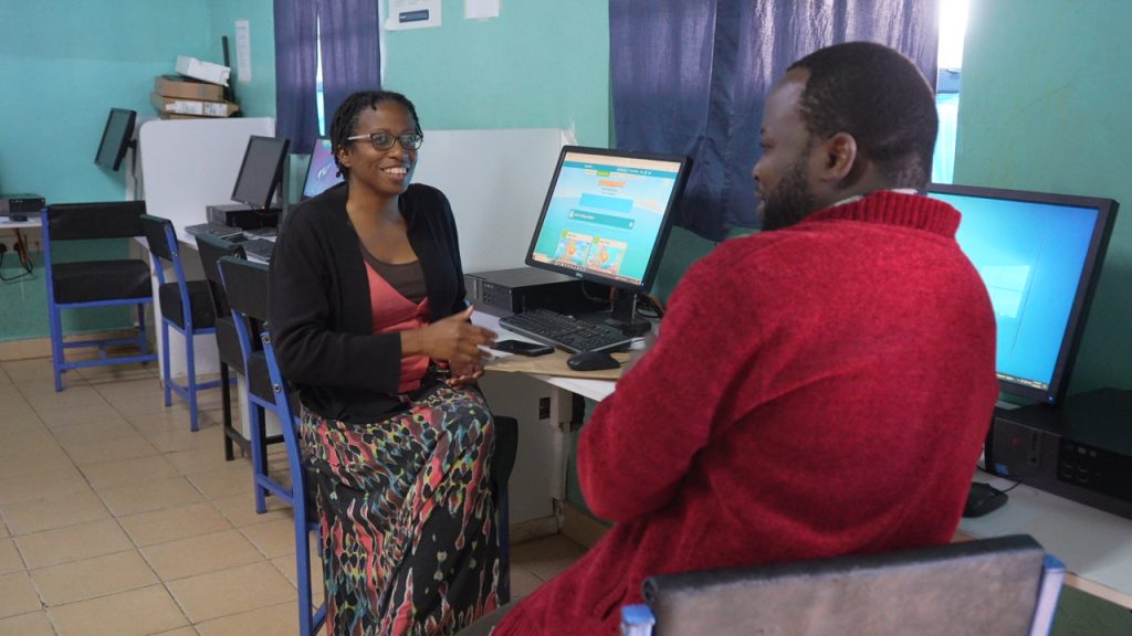 A woman and man talk beside desktop computers in a turquoise-walled computer lab.