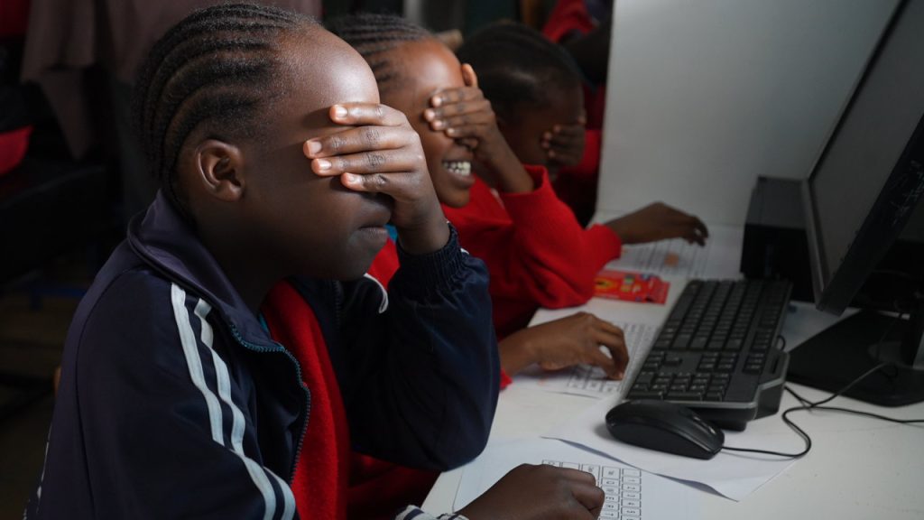 Schoolchildren seated at computers cover their eyes with their hands during a playful classroom activity.