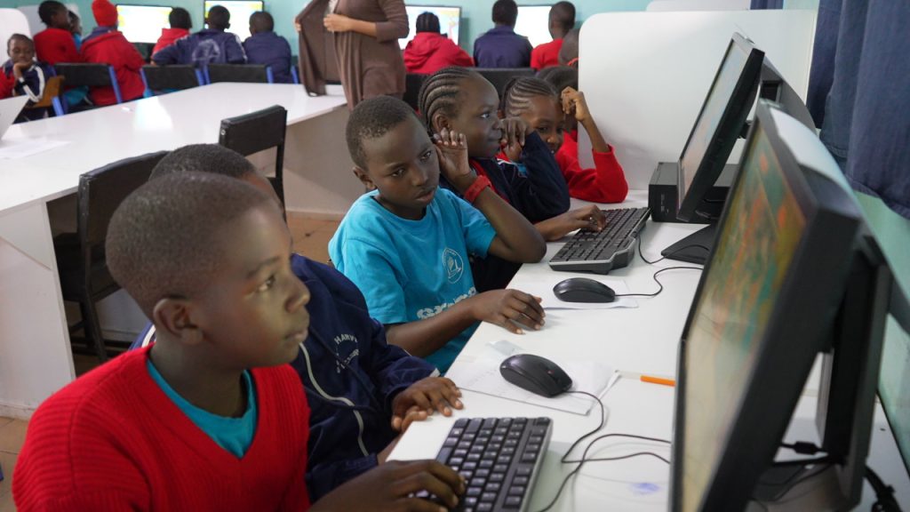 A group of schoolchildren sit at desktop computers, concentrating on activities displayed on their screens.