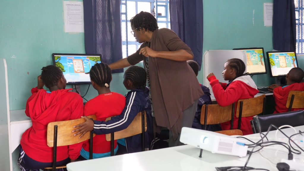 An instructor points toward a computer screen while guiding a group of children seated at desktop workstations.
