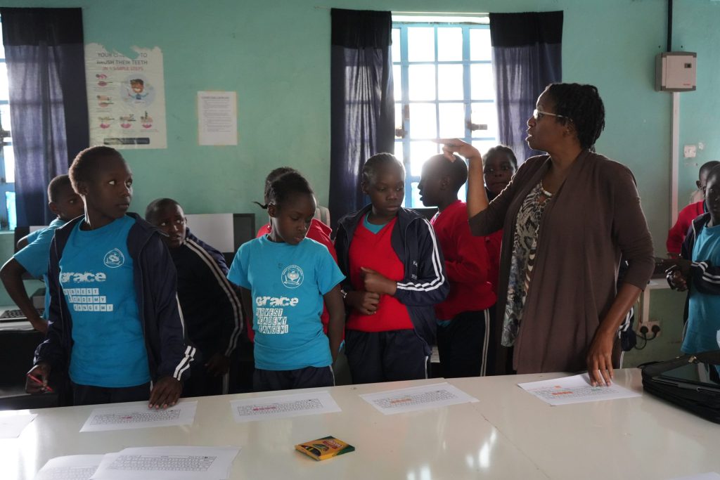 An instructor gestures while schoolchildren stand around a table with printed keyboard worksheets.