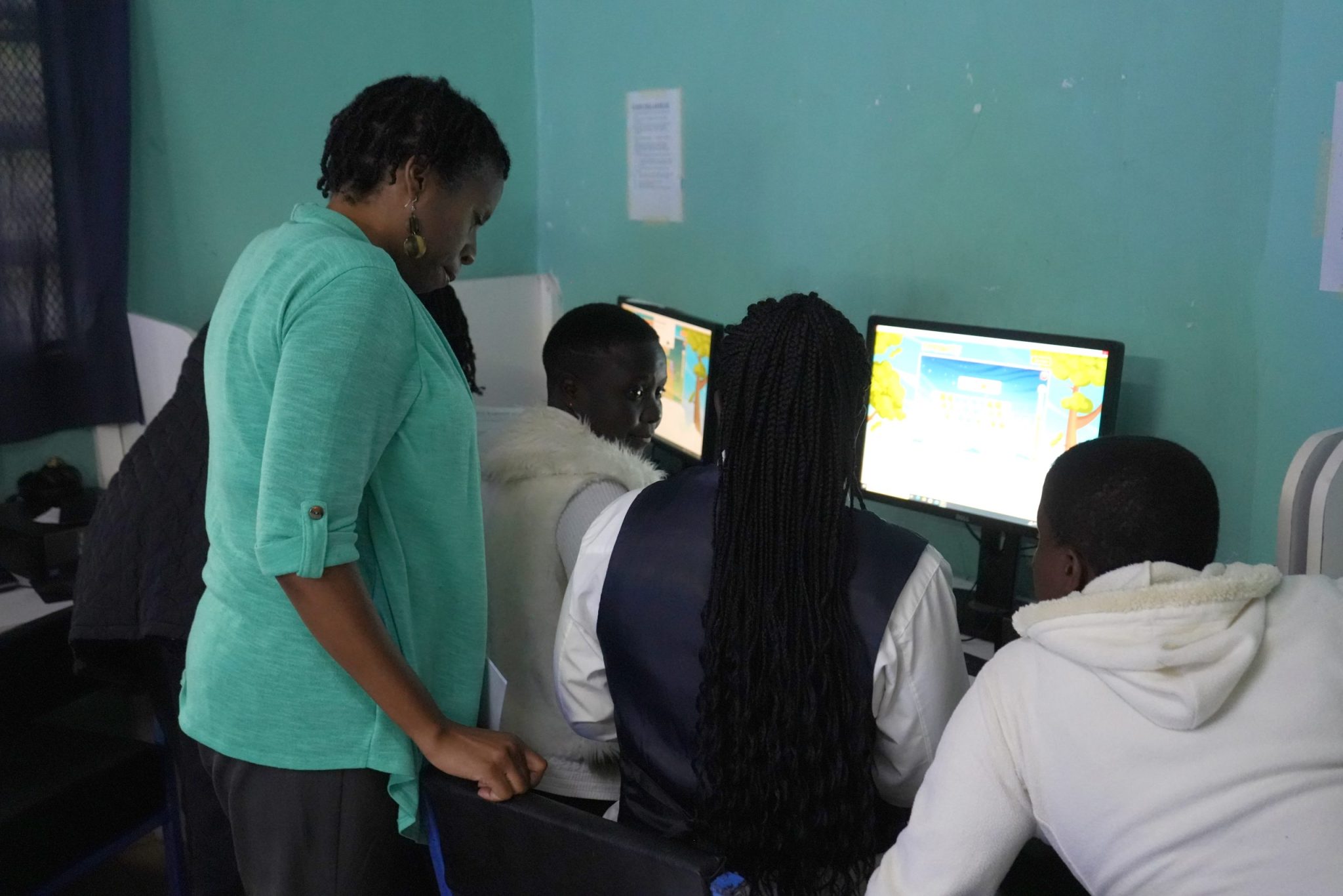 An instructor stands beside three participants seated at desktop computers in a classroom computer lab.