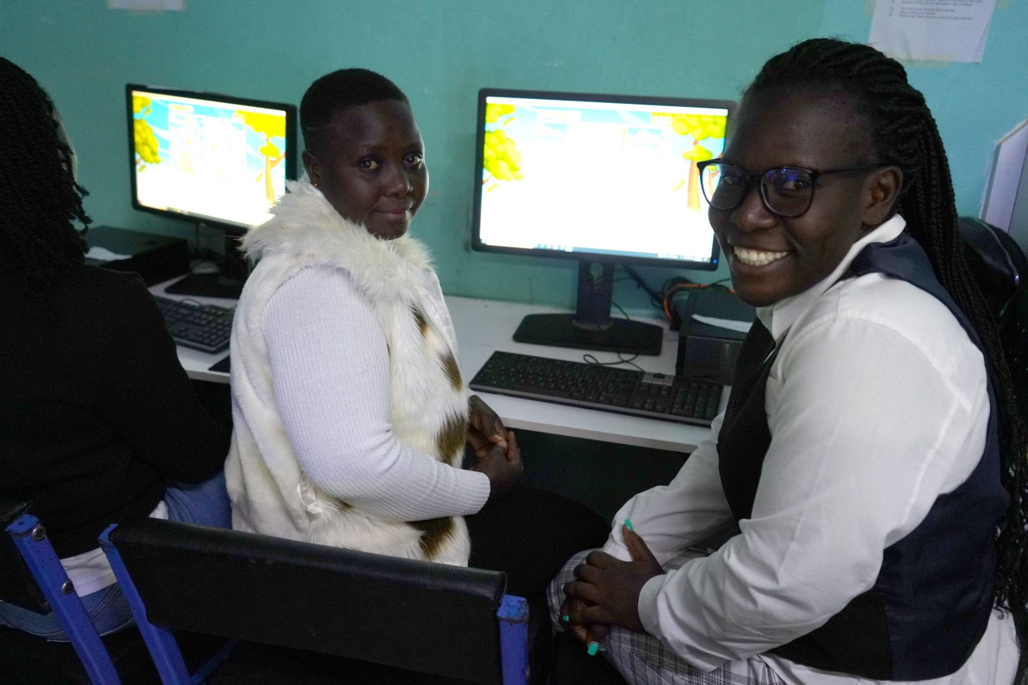 Two participants seated at computer stations turn toward the camera, one smiling, during a computer training session.