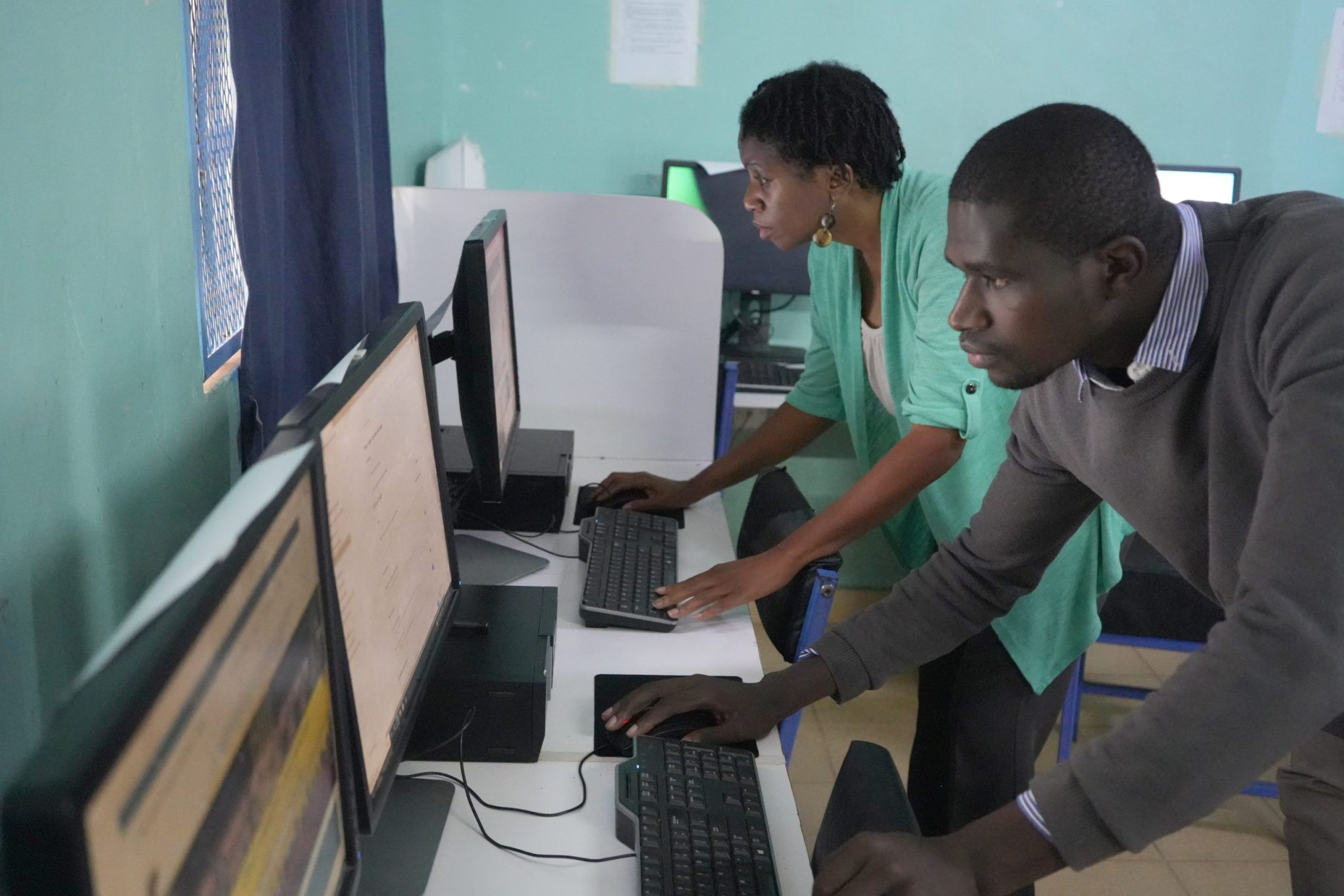 Two instructors lean toward desktop computers while working at adjacent stations in a computer lab.