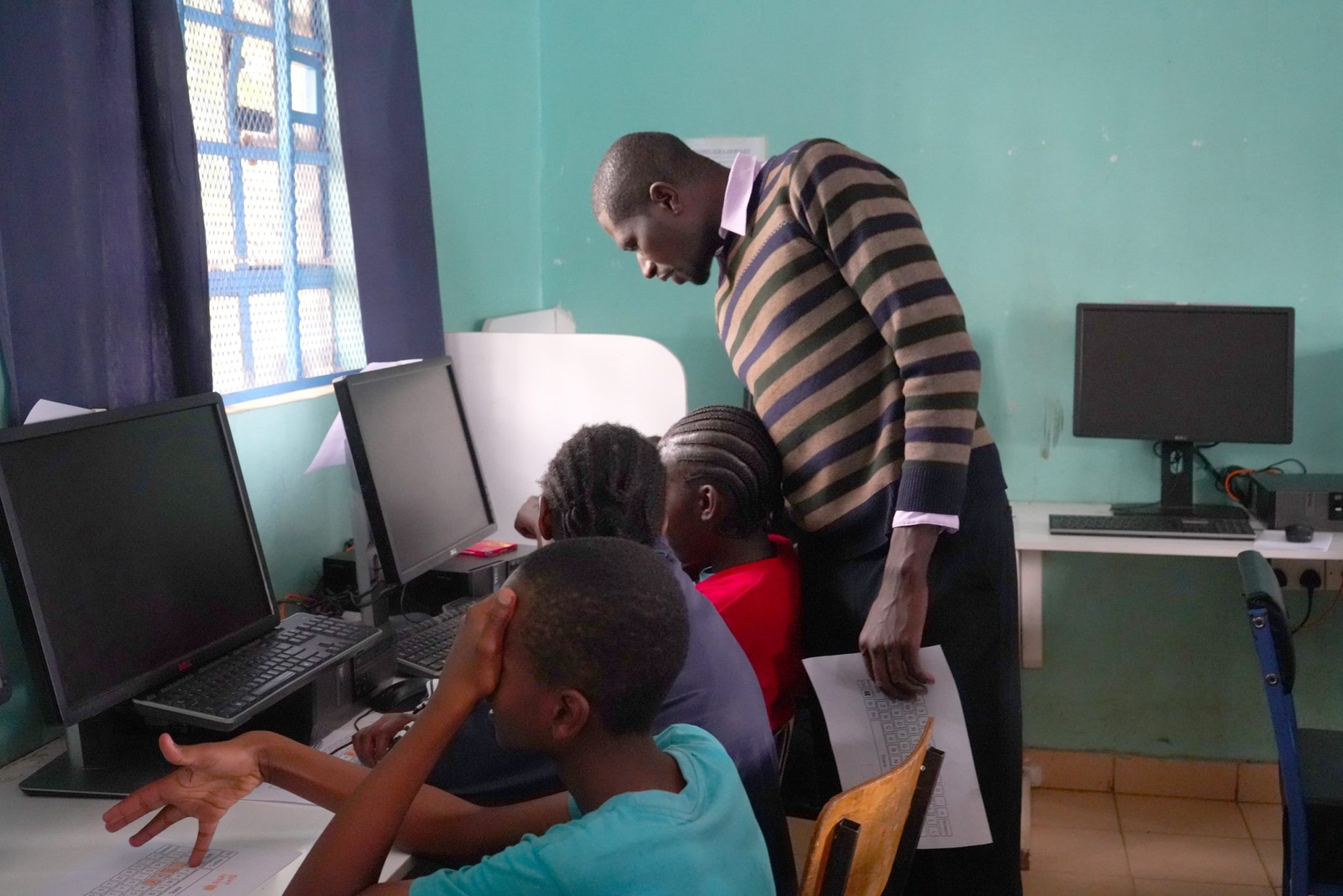 An instructor stands beside three seated learners working at computers and with printed worksheets in a computer lab.