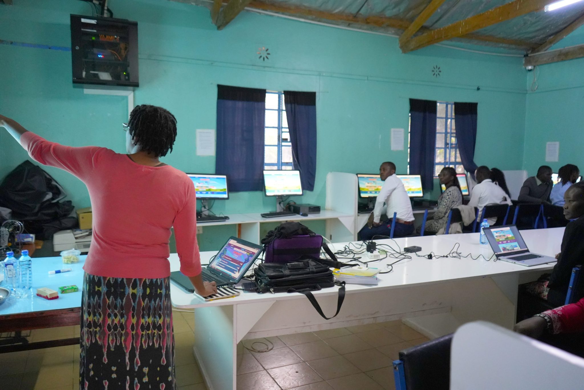 An instructor stands at the front of a computer lab addressing participants seated around rows of desktop computers.