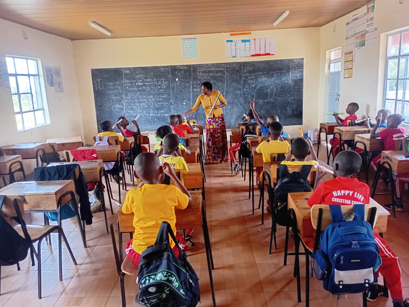 Students at Happy Life Christian School in Juja Farm, Kenya, practice colorful recorders at their desks while Kennesha leads the music lesson from the front of the classroom.