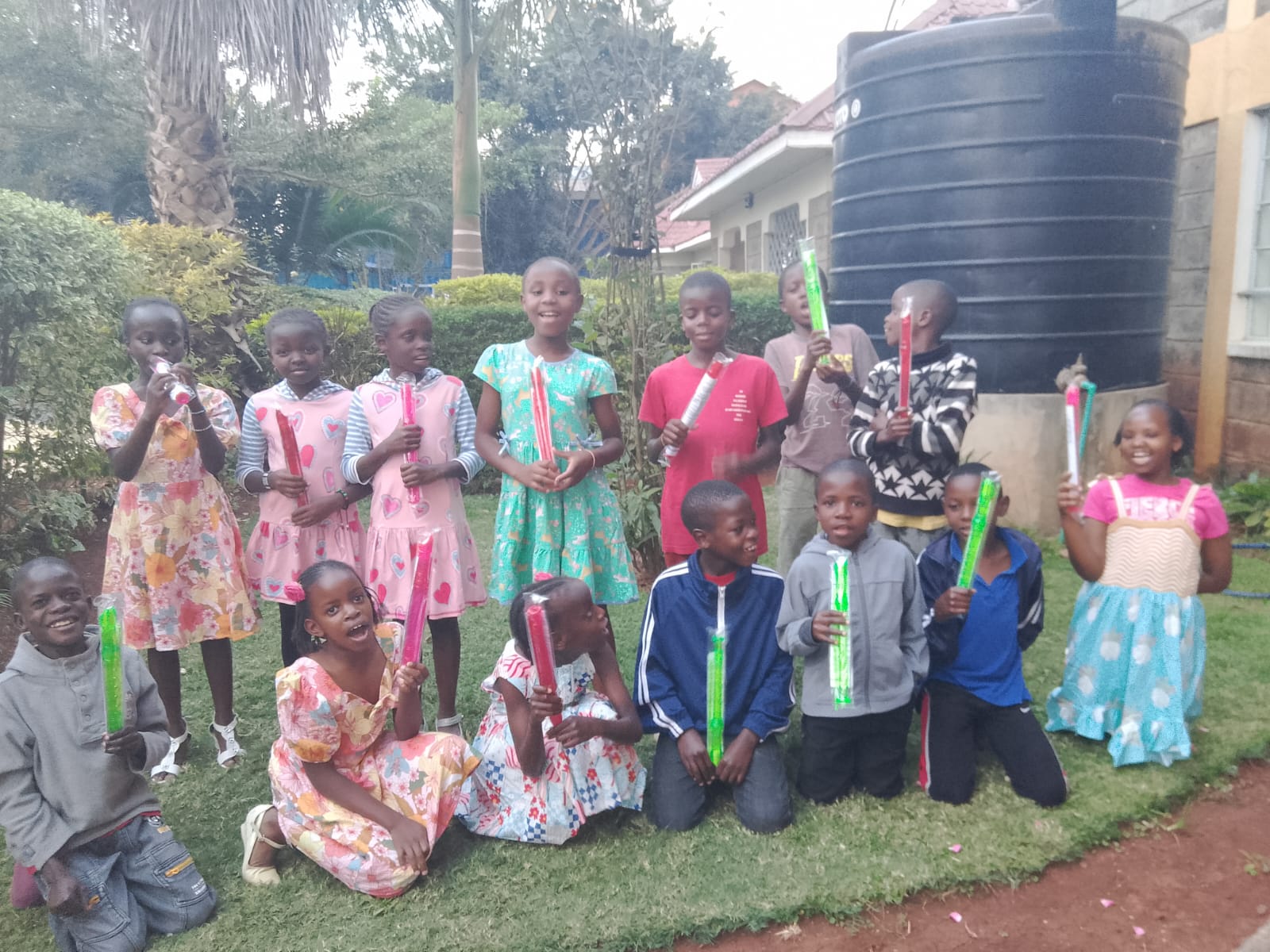 Children at Happy Life in Juja Farm, Kenya, gather outdoors on the grass while holding colorful musical tubes during a group music activity.