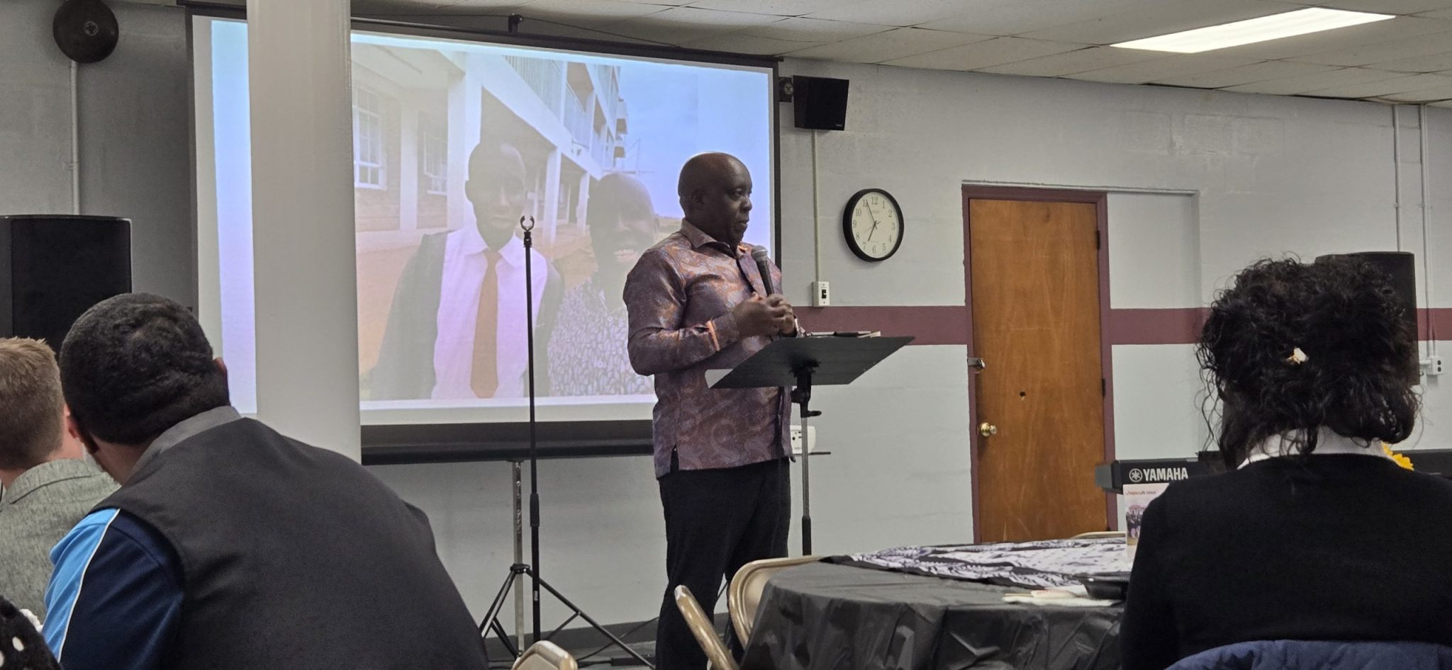 Bishop Peter addressing guests during a Happy Life Mission-Kenya presentation with a projected photograph behind him