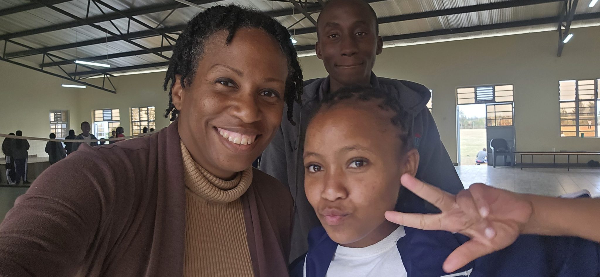 Kennesha taking a selfie with two students, Faith and Jonathan, inside a large school activity room.
