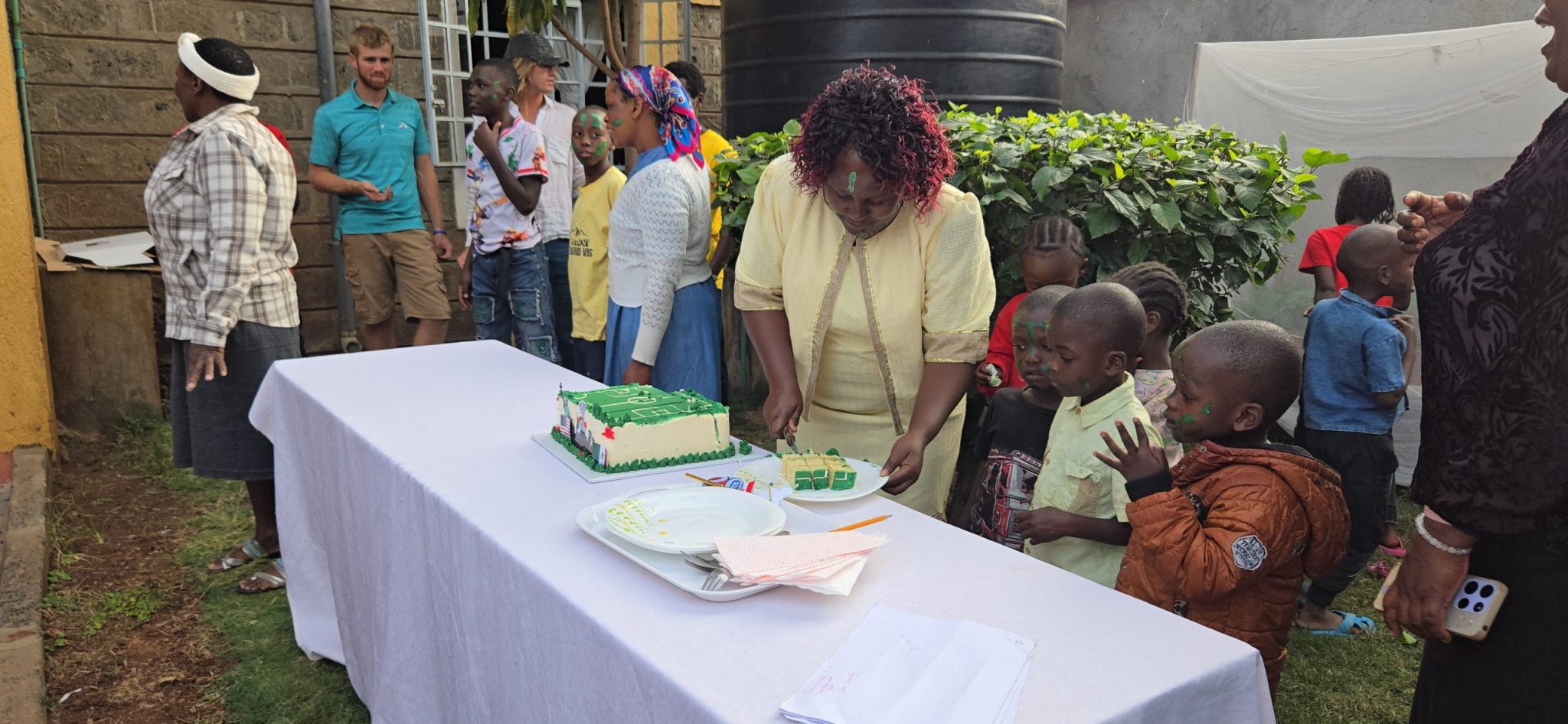 Mom Tere cutting a green soccer-themed cake while children gather nearby outdoors.