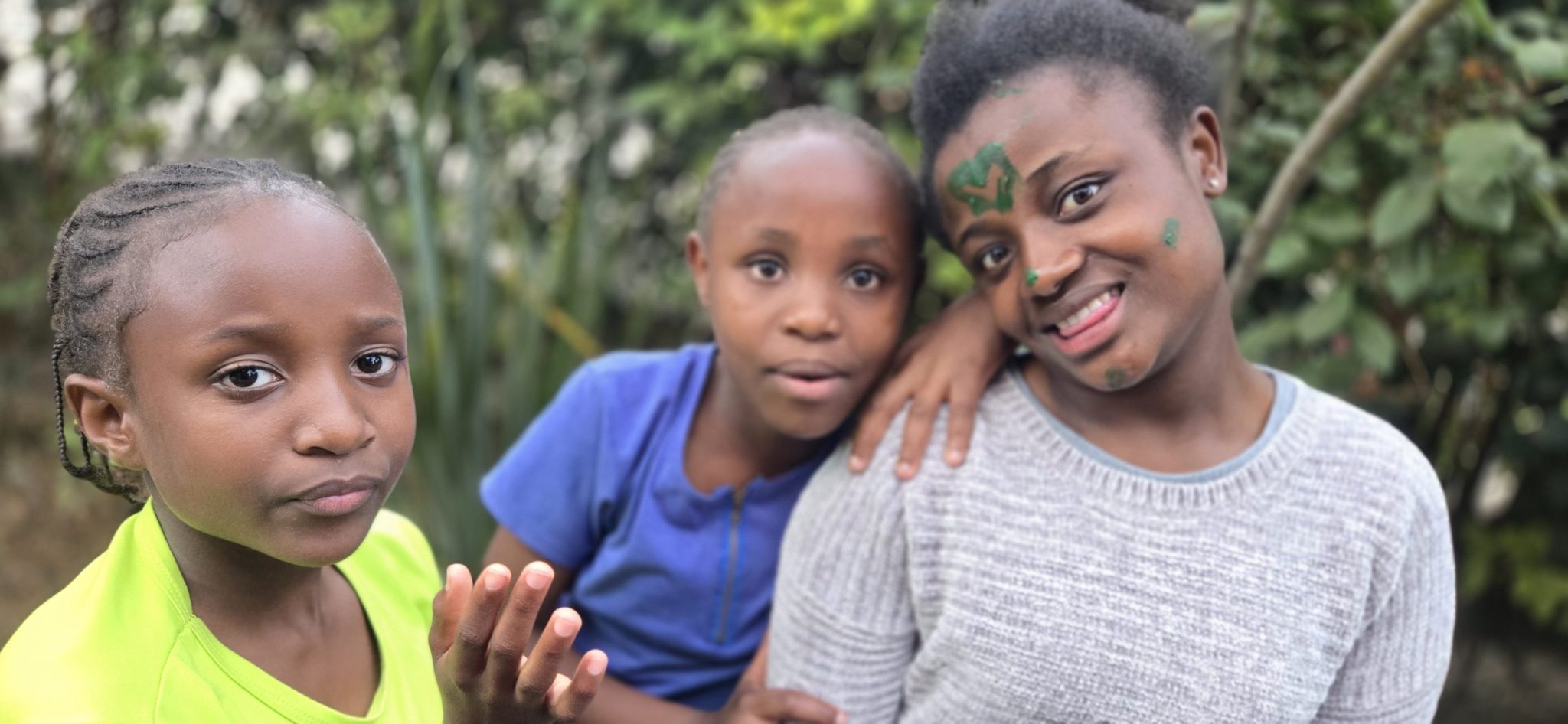 Three girls, Hadassah, Shiko, and Leah, posing together outdoors at Happy Life, including one with green face paint.