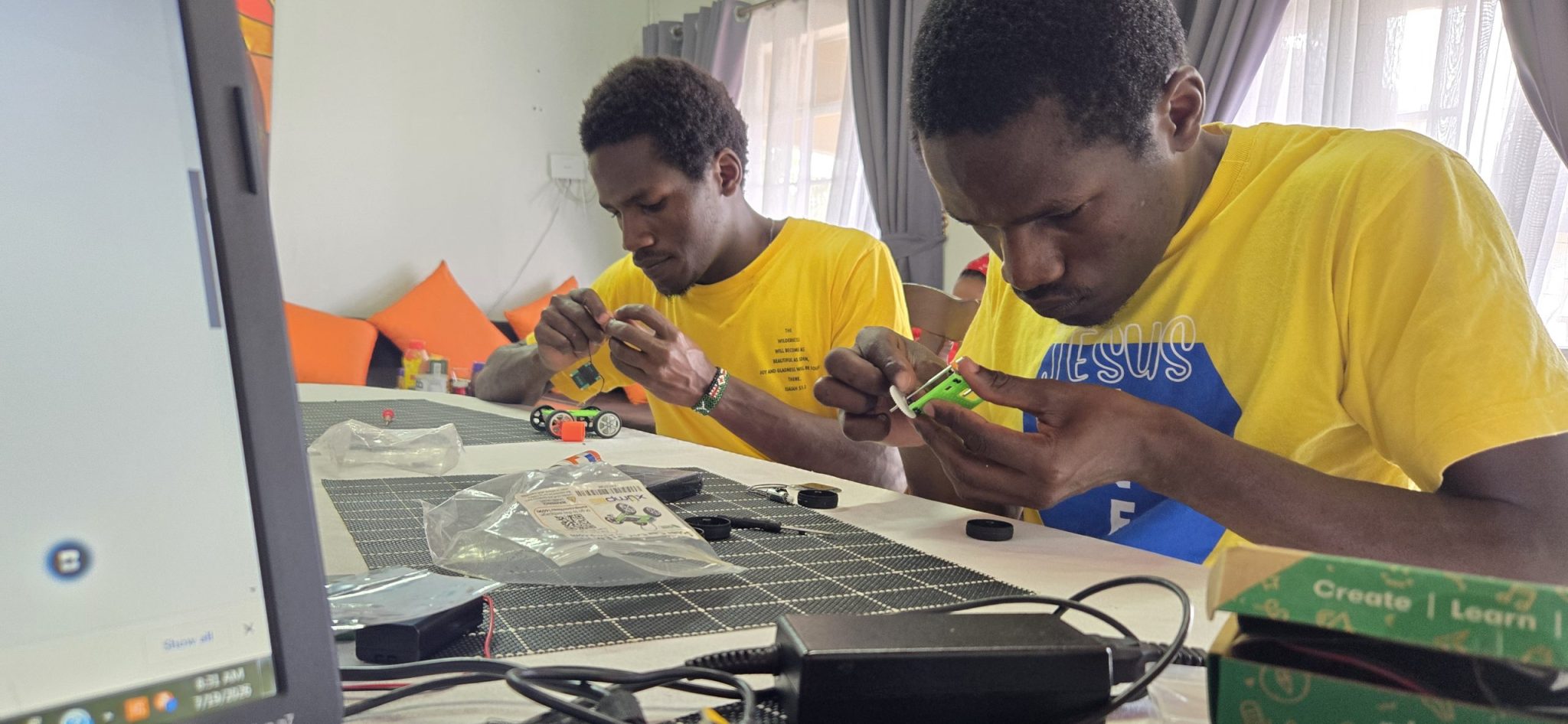 Two young men, Peter and Dominic, assembling small electronics and vehicle kits beside a laptop.