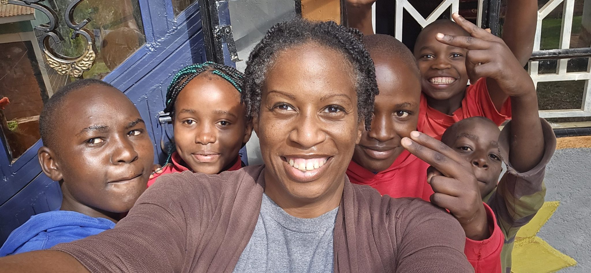 Kennesha, smiling, takes a close-up group selfie with five children gathered around her outside a building entrance. Several children smile or make playful hand gestures toward the camera.