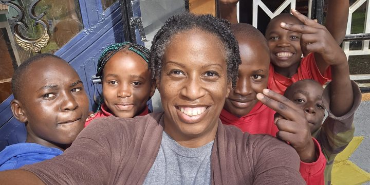 Kennesha, smiling, takes a close-up group selfie with five children gathered around her outside a building entrance. Several children smile or make playful hand gestures toward the camera.
