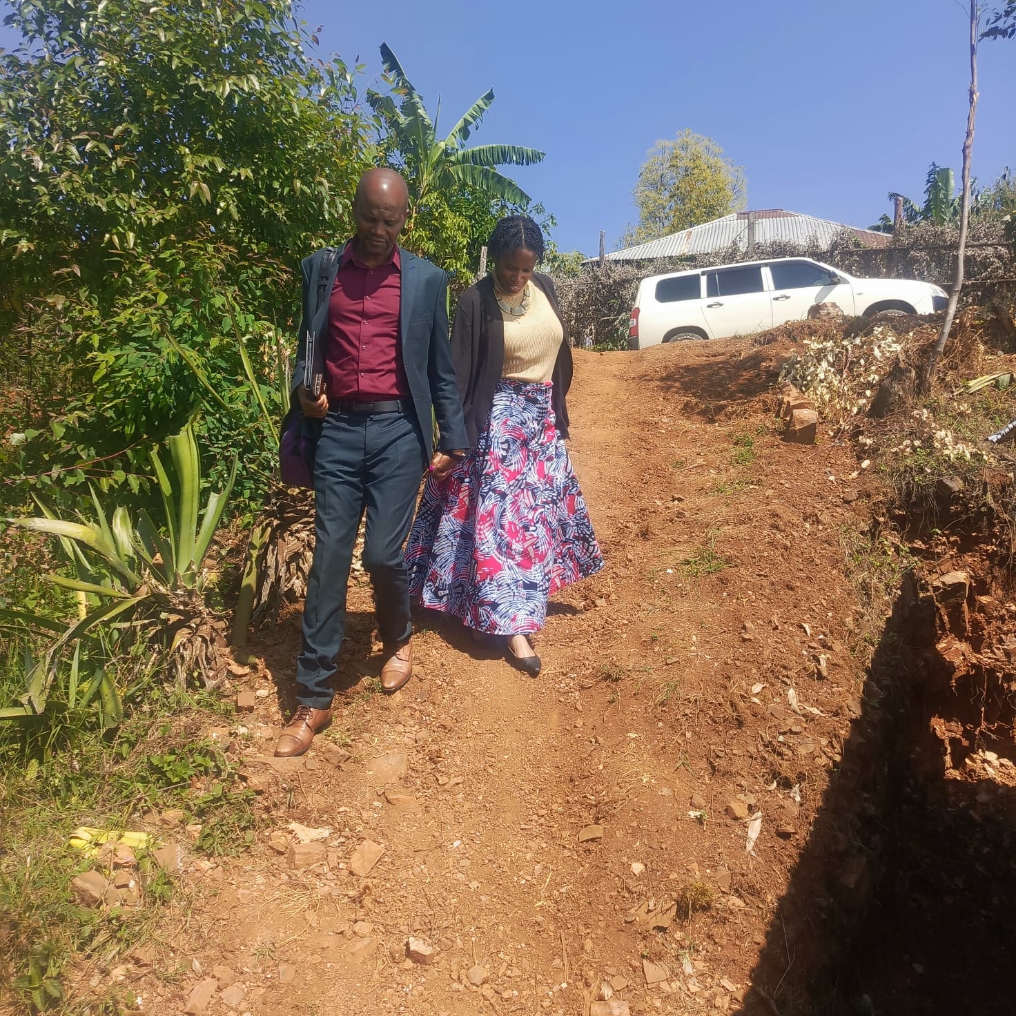 A man in a dark suit and a woman in a long patterned skirt walk carefully down a steep dirt path, holding hands. Lush vegetation, banana plants, a house, and a white SUV appear behind them under a clear blue sky.