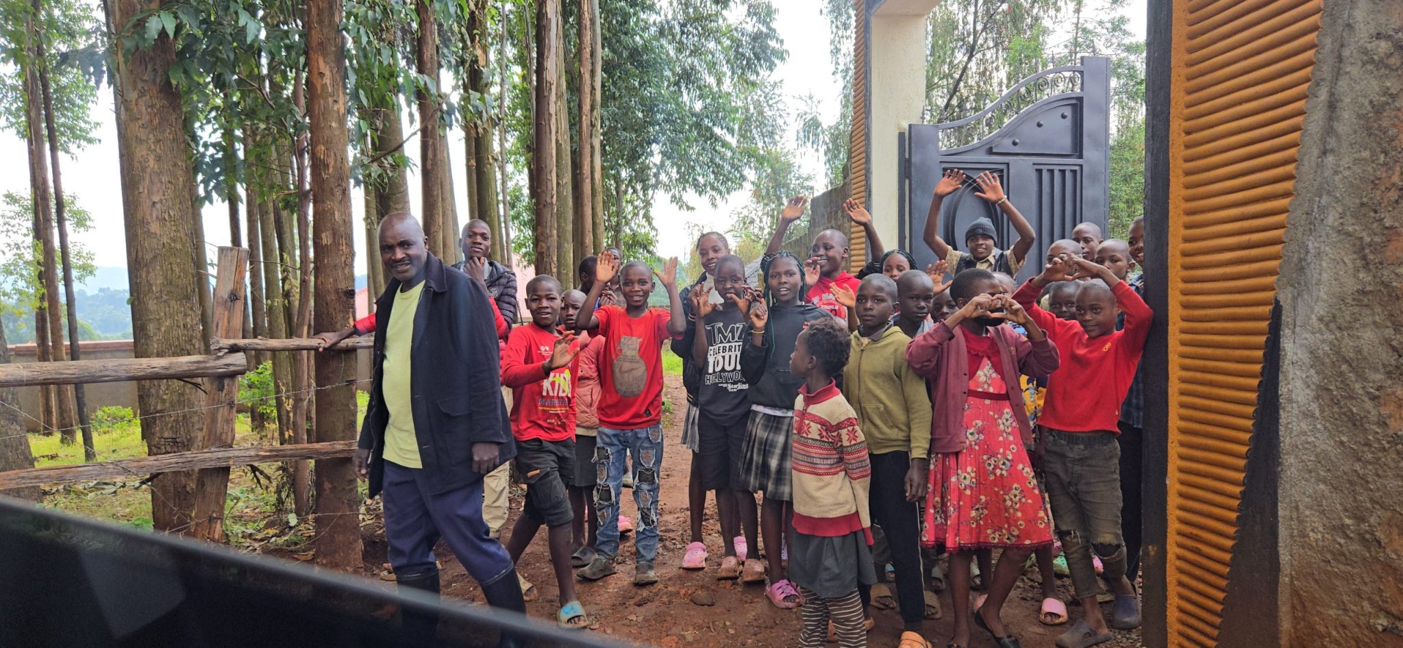 Children waving beside an adult near the entrance of a property surrounded by trees in Kisii, Kenya.